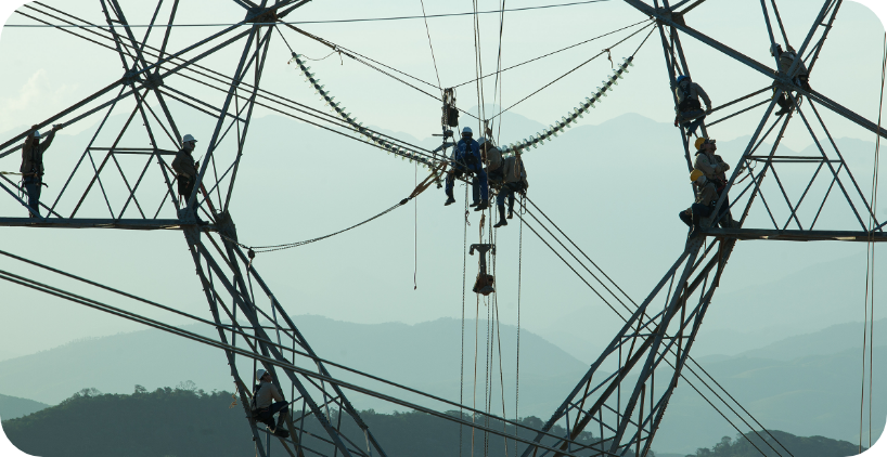 Foto mostra, em primeiro plano, nove empregados trabalhando na manutenção de emergência de uma torre de transmissão. Ao fundo, um horizonte com uma mata verde e montanhas arredondadas. Fim da descrição.