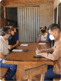 Foto mostra empregados de Furnas, um homem e uma mulher, realizando pesquisa em campo com um membro da comunidade em Minas Gerais. No total, quatro pessoas estão sentadas à mesa de madeira, usando máscaras de proteção contra a Covid-19, e com papéis brancos à frente de cada um.